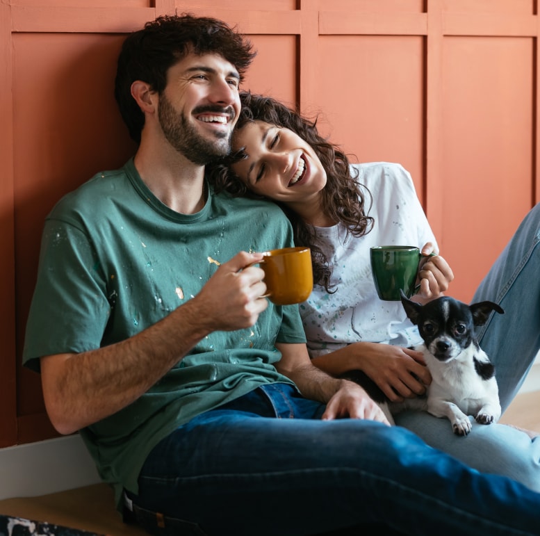 A smiling couple each holding a coffee mug sit together on the floor with their dog.
